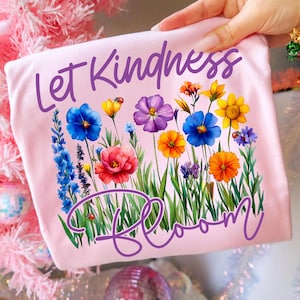 May include: A light pink t-shirt featuring the words "Let Kindness Bloom" in a purple script. The shirt has a colorful floral design with blue, yellow, orange, and purple flowers.