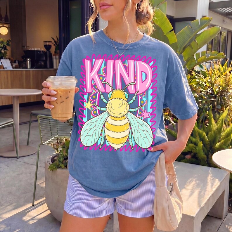 May include: Blue t-shirt with a graphic of a bee and the word "KIND" in pink and blue. The bee is yellow and white with blue wings. The shirt is paired with blue and white striped shorts.