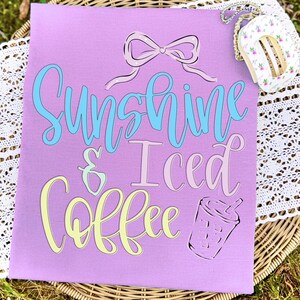 Op de afbeelding: Een lila t-shirt met de tekst "Sunshine & Iced Coffee" in een speels lettertype. De tekst is in lichtblauw, roze en geel. Een strik en een koffiekopje staan ook op de shirt. Een klein item met bloemenpatroon staat in de rechterbovenhoek.
