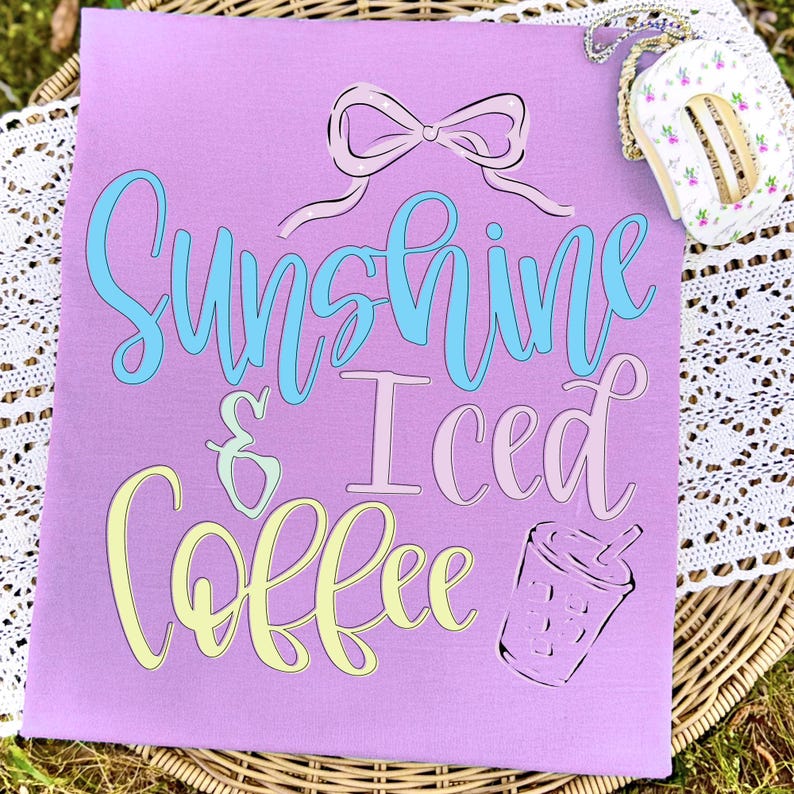 Op de afbeelding: Een lila t-shirt met de tekst "Sunshine & Iced Coffee" in een speels lettertype. De tekst is in lichtblauw, roze en geel. Een strik en een koffiekopje staan ook op de shirt. Een klein item met bloemenpatroon staat in de rechterbovenhoek.