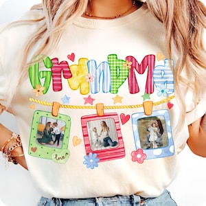 May include: Cream-colored t-shirt with the word "Grandma" in colorful, patterned letters. Below, three framed photos hang from a rope, decorated with hearts and flowers. The photos appear to show family members.