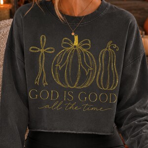 K&ouml;nnte beinhalten: Ein anthrazitfarbenes Langarm-Sweatshirt mit einer goldglitzernden Grafik, die ein Kreuz, einen runden K&uuml;rbis mit Schleife und einen l&auml;nglichen K&uuml;rbis zeigt. Der Text "GOD IS GOOD all the time" steht in goldglitzernden Buchstaben auf der Brust.