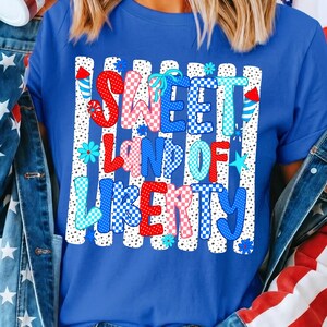 May include: A royal blue t-shirt featuring a graphic print with the text SWEET LAND OF LIBERTY in red, white, and blue checkered and polka dot patterns. The design includes small firework and flower illustrations over vertical white stripes.