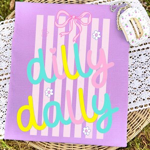 May include: A lilac canvas featuring vertical pink and white stripes. The words "dilly dally" are written in a playful font with yellow, pink, and turquoise colours. A pink bow and small white flowers decorate the canvas.