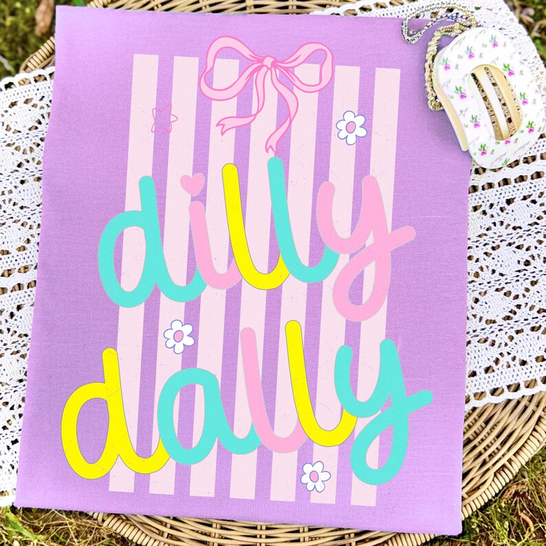 May include: A lilac canvas featuring vertical pink and white stripes. The words "dilly dally" are written in a playful font with yellow, pink, and turquoise colours. A pink bow and small white flowers decorate the canvas.