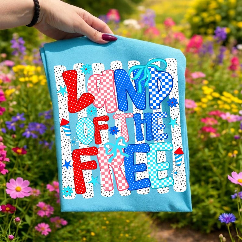 Puede incluir: Una camiseta azul claro con el texto LAND OF THE FREE en una fuente llamativa y multicolor con patrones de lunares y cuadros en rojo, blanco y azul. El dise&ntilde;o incluye peque&ntilde;as estrellas azules y gr&aacute;ficos de fuegos artificiales.