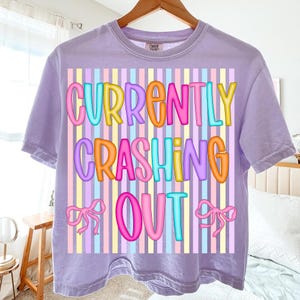 Pode incluir: Uma camiseta lil&aacute;s com a frase "CURRENTLY CRASHING OUT" em letras coloridas. O texto est&aacute; sobre um fundo listrado de cores pastel, com la&ccedil;os rosa em ambos os lados da palavra "OUT".
