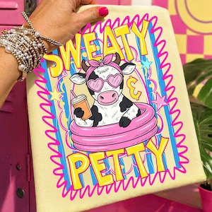 May include: A light yellow towel with the words "SWEATY PETTY" in yellow text with a pink and blue outline. The towel features a cartoon cow wearing heart-shaped sunglasses and a pink headband, floating in a pink inflatable ring, holding a drink.