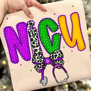 May include: A light beige shirt with the word "NICU" in large, colorful letters. The letters are purple, green, and yellow, with a leopard print bow and a purple stethoscope. The design includes white polka dots and decorative purple trim.