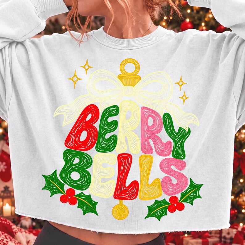 May include: A white long-sleeve cropped sweatshirt featuring a graphic of a large bell with a bow. The text BERRY BELLS is written in a colorful, stylized font across the bell. Green holly leaves with red berries are at the bottom of the design.