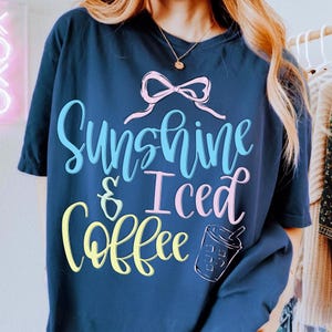 Op de afbeelding: Marineblauwe t-shirt met de tekst "Sunshine & Iced Coffee" in een speels lettertype. Het ontwerp bevat een roze strik en een afbeelding van een ijskoffie. Het shirt is een casual, comfortabele stijl.