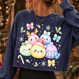 May include: A navy blue long-sleeve cropped sweatshirt featuring a graphic print of five colourful, smiling dumpling characters. The dumplings are decorated with a Santa hat, a blue scarf, and reindeer antlers, surrounded by bows, snowflakes, and holly.