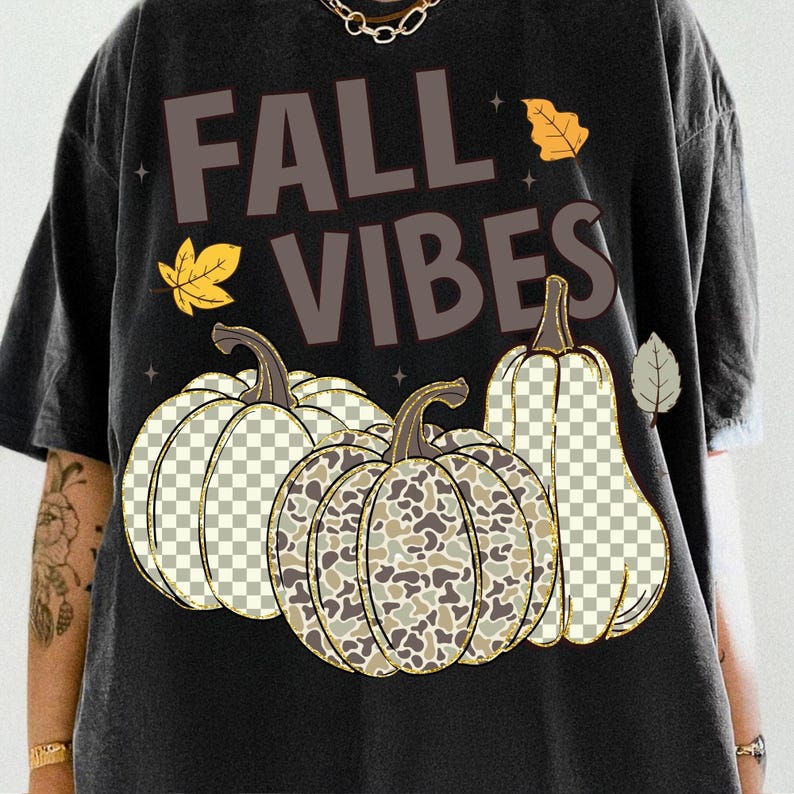 Puede incluir: Una camiseta negra con el texto FALL VIBES en letras marrones. El gr&aacute;fico incluye tres calabazas con patrones de cuadros y camuflaje, acentuadas por peque&ntilde;as hojas amarillas y grises y diminutas formas de estrellas dispersas por el dise&ntilde;o.