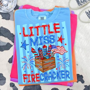 May include: A light blue t-shirt with the words "LITTLE MISS FIRECRACKER" in red and blue. The shirt features a graphic of fireworks, stars, and an American flag. The shirt is folded on top of other colorful shirts.