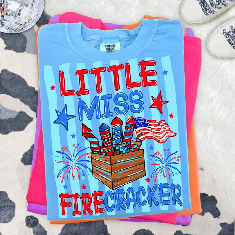 May include: A light blue t-shirt with the words "LITTLE MISS FIRECRACKER" in red and blue. The shirt features a graphic of fireworks, stars, and an American flag. The shirt is folded on top of other colorful shirts.