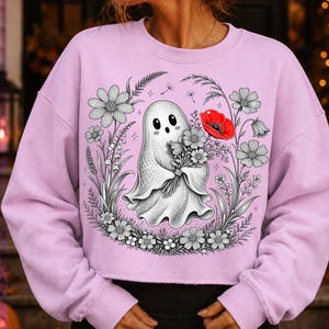 K&ouml;nnte beinhalten: Ein hellviolettes Langarm-Sweatshirt mit einer zentralen Grafik eines wei&szlig;en Geistes, der einen Blumenstrau&szlig; h&auml;lt. Der Geist ist von einer kreisf&ouml;rmigen Anordnung aus schwarz-wei&szlig;en Strichzeichnungen von Blumen umgeben, inklusive einer roten Mohnblume.