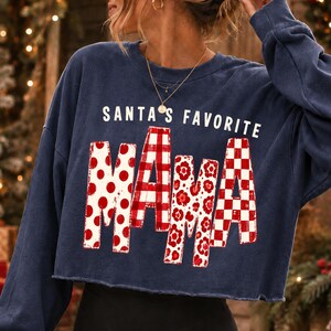 K&ouml;nnte beinhalten: Ein marineblaues, lang&auml;rmeliges, bauchfreies Sweatshirt mit dem wei&szlig;en Text SANTA'S FAVORITE &uuml;ber dem Wort MAMA. Die Buchstaben in MAMA sind mit verschiedenen rot-wei&szlig;en Mustern gef&uuml;llt, darunter Punkte, Karos, Blumen und ein Schachbrettmuster.