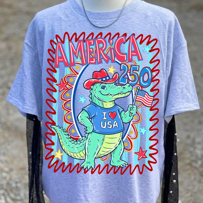 Puede incluir: Camiseta gris claro con un cocodrilo de dibujos animados que lleva un sombrero de vaquero y una camiseta azul con "I ❤️ USA". El dise&ntilde;o incluye el texto "AMERICA 250" en rojo y azul, con estrellas y una bandera americana ondeando.