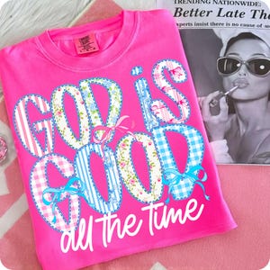 Könnte beinhalten: Ein leuchtend pinkfarbenes T-Shirt mit dem Aufdruck "GOD IS GOOD all the time". Die Buchstaben sind mit floralen, gestreiften und Vichy-Mustern gestaltet und mit blauen Schleifen verziert. Das T-Shirt liegt auf einer rosa Oberfläche.
