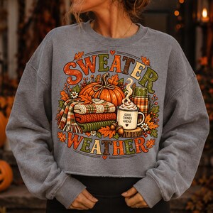 Puede incluir: Una sudadera gris jaspeado de cuello redondo con un gr&aacute;fico colorido que incluye el texto "SWEATER WEATHER". El dise&ntilde;o muestra una calabaza naranja grande, una pila de jers&eacute;is doblados, un termo y una taza con el texto "GOOD THINGS AHEAD".