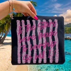 May include: Black square tote bag with pink vertical stripes and the text "If The Stars Were Made To Worship So Will I." The bag is held up against a tropical beach background.
