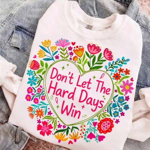 K&ouml;nnte beinhalten: Wei&szlig;es Sweatshirt mit einem farbenfrohen Blumendesign, das eine herzf&ouml;rmige Grafik umgibt. Das Herz enth&auml;lt den Text "Don't Let The Hard Days Win" in Rosa. Die Blumen sind in Rosa-, Gelb-, Blau- und Oranget&ouml;nen gehalten.