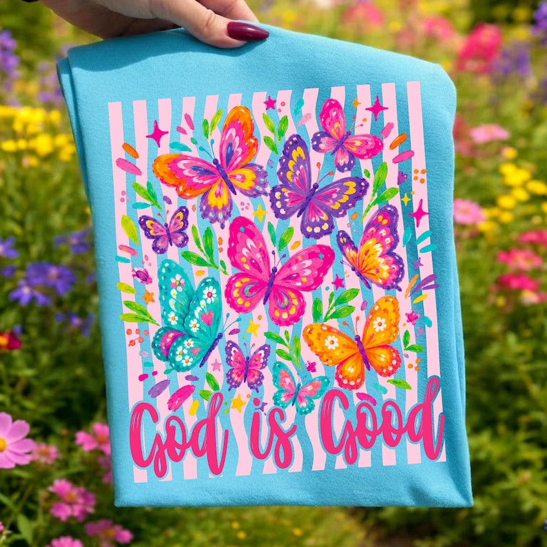 May include: A light blue t-shirt featuring a graphic print of multiple colorful butterflies in shades of pink, orange, purple, and teal. The design includes vertical light pink stripes, small star accents, and the text God is Good in pink script.