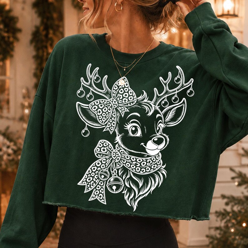 May include: A dark green long-sleeve cropped sweatshirt featuring a white graphic print of a reindeer head. The reindeer has a leopard print bow between its antlers and a matching leopard print scarf around its neck with two hanging jingle bells.
