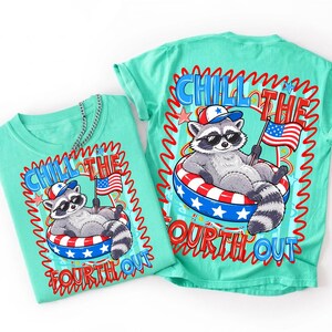 Op de afbeelding: Twee turquoise t-shirts met een afbeelding van een wasbeer met een zonnebril en een patriotische hoed, liggend in een rood, wit en blauw zwemband. De tekst "Chill the Fourth Out" staat op de shirts.