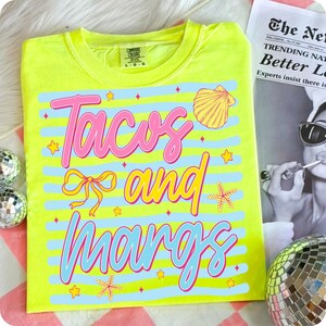 May include: A neon yellow t-shirt with the words "Tacos and Margs" in pink and blue, decorated with a seashell, bow, starfish, and stars. The shirt is a Comfort Colors brand, size L-G.
