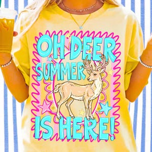 K&ouml;nnte beinhalten: Gelbes T-Shirt mit dem Text "OH DEER SUMMER IS HERE!" in T&uuml;rkis, umrandet von Pink. Eine Illustration eines beigen Hirsches befindet sich darunter, umgeben von rosa Sternen und Wellenlinien.
