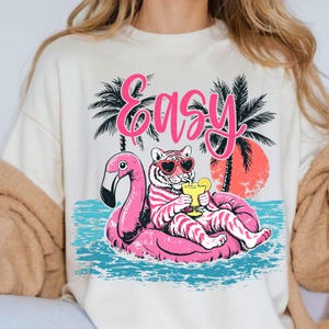 K&ouml;nnte beinhalten: Cremefarbenes Sweatshirt mit einem Aufdruck eines Tigers mit herzf&ouml;rmiger Sonnenbrille, der auf einem rosa Flamingo-Schwimmer entspannt. Das Wort "Easy" steht in rosa Schrift &uuml;ber dem Tiger, mit Palmen und einem Sonnenuntergang im Hintergrund.