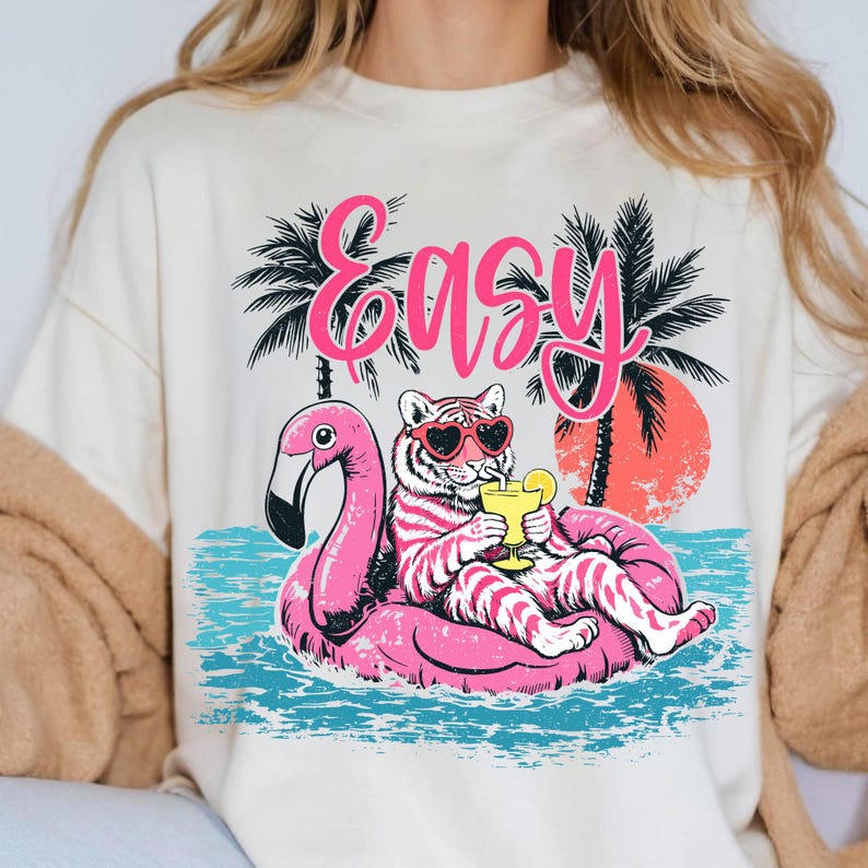 K&ouml;nnte beinhalten: Cremefarbenes Sweatshirt mit einem Aufdruck eines Tigers mit herzf&ouml;rmiger Sonnenbrille, der auf einem rosa Flamingo-Schwimmer entspannt. Das Wort "Easy" steht in rosa Schrift &uuml;ber dem Tiger, mit Palmen und einem Sonnenuntergang im Hintergrund.