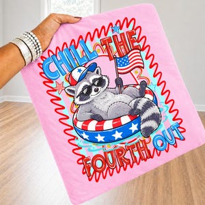 Op de afbeelding: Een roze deken met een cartoon wasbeer met een zonnebril en een patriotische hoed, liggend in een zwemband met een Amerikaanse vlag. De deken heeft de tekst "Chill the Fourth Out" in rood en blauw.
