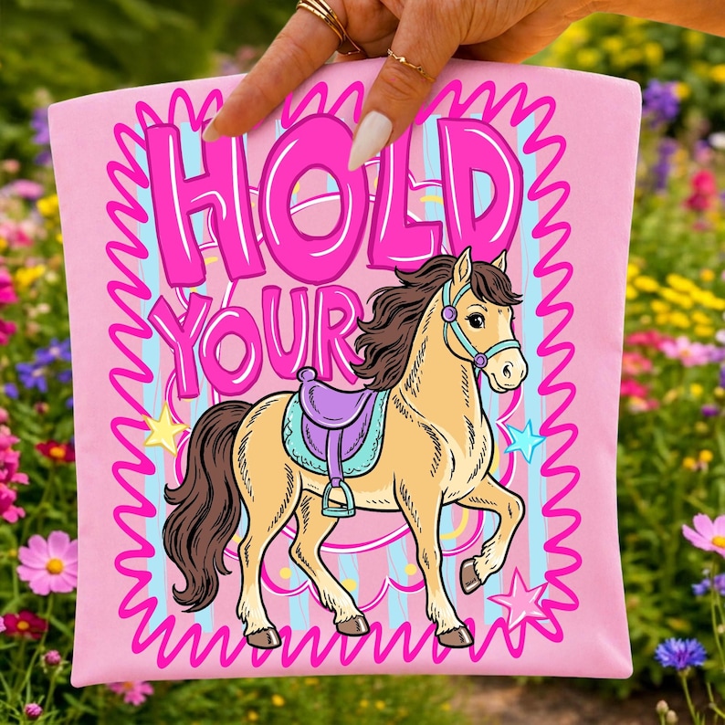 May include: Pink t-shirt with the words "HOLD YOUR" in large pink letters and a cartoon horse illustration. The horse is tan with a purple saddle and blue bridle. Stars and a wavy pink border surround the design.