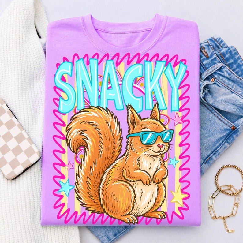 May include: A lilac-colored t-shirt featuring a cartoon squirrel wearing blue sunglasses. The word "SNACKY" is displayed in large, turquoise letters above the squirrel. The design includes pink, yellow, and blue accents.