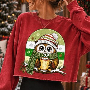 May include: A red long-sleeve cropped sweatshirt featuring a graphic of a brown owl wearing a red, white, and green striped knit hat and a green cable-knit scarf. The owl holds a yellow mug with white steam, set against a green background.