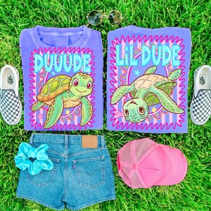 May include: Two lilac t-shirts with turtle graphics and the words "DUUUDE" and "LIL DUDE". The image also shows denim shorts, a pink trucker hat, a blue scrunchie, sunglasses, and checkered slip-on shoes, all on a green grassy surface.