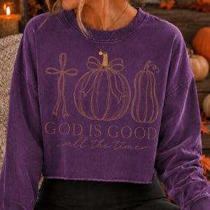 K&ouml;nnte beinhalten: Ein violettes Langarm-Sweatshirt mit Rundhalsausschnitt und offener Saumkante. Auf der Vorderseite befindet sich eine goldfarbene Strichzeichnung eines Kreuzes, eines K&uuml;rbisses mit Schleife und einer K&uuml;rbisfrucht. Der Text lautet "GOD IS GOOD all the time".