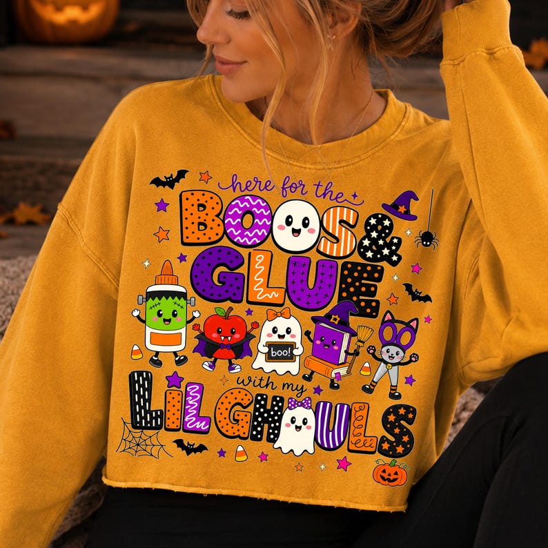 K&ouml;nnte beinhalten: Ein senfgelbes bauchfreies Sweatshirt mit einem Grafikdruck und dem Text "here for the BOOS & GLUE with my LIL GHOULS". Das Design zeigt Zeichentrickfiguren wie eine Klebeflasche, einen Apfel, ein Gespenst, ein Buch und eine Katze sowie Flederm&auml;use.