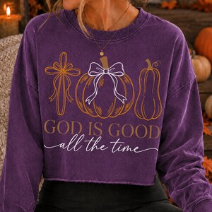 Puede incluir: Una sudadera morada de manga larga con cuello redondo que presenta un estampado gr&aacute;fico de una cruz, una calabaza con un lazo blanco y una calabaza alargada. El texto dice "GOD IS GOOD" en letras de molde y "all the time" en cursiva.