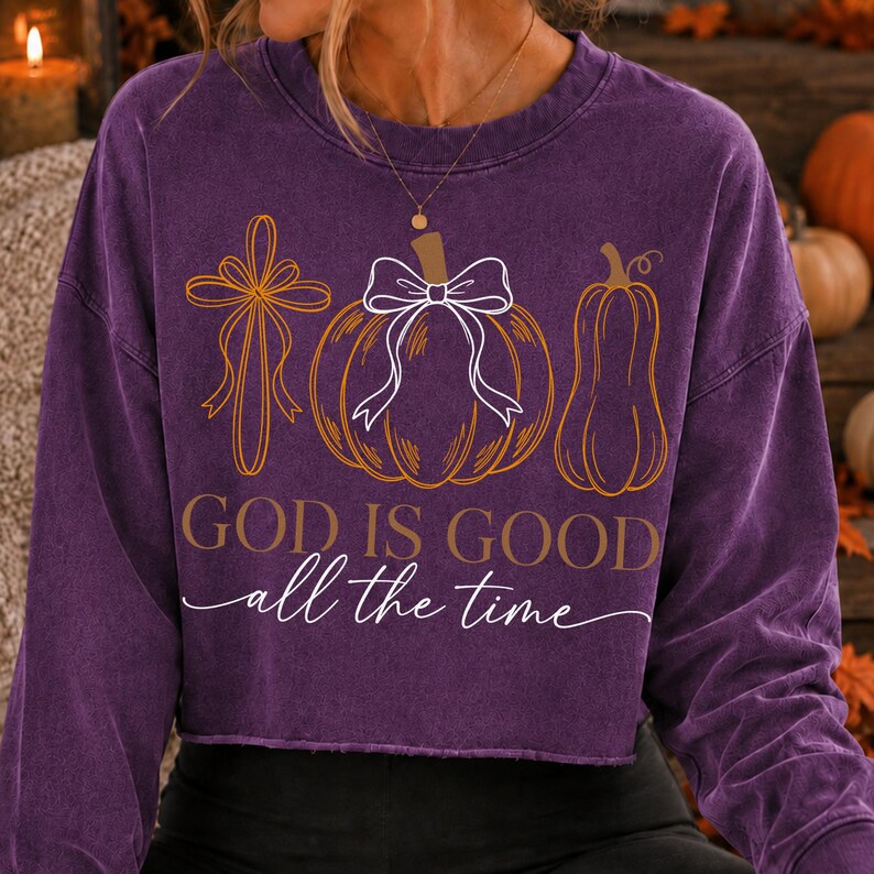 Puede incluir: Una sudadera morada de manga larga con cuello redondo que presenta un estampado gr&aacute;fico de una cruz, una calabaza con un lazo blanco y una calabaza alargada. El texto dice "GOD IS GOOD" en letras de molde y "all the time" en cursiva.