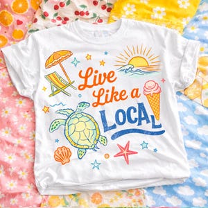 K&ouml;nnte beinhalten: Wei&szlig;es T-Shirt mit umgekrempelten &Auml;rmeln und einem farbenfrohen Strandmotiv. Das Design zeigt eine Schildkr&ouml;te, ein Eis, einen Strandstuhl und den Text "Live Like a Local". Das Shirt ist mit Sternen, einer Muschel und einem Seestern verziert.