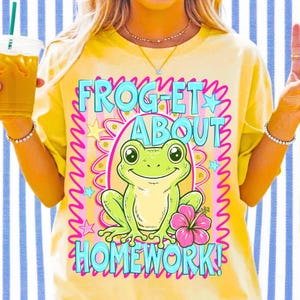 K&ouml;nnte beinhalten: Gelbes T-Shirt mit einem Frosch-Cartoon und dem Text "FROGET ABOUT HOMEWORK!". Der Frosch ist gr&uuml;n mit einer rosa Blume. Der Text ist blau und rosa. Eine Person tr&auml;gt das T-Shirt und h&auml;lt ein Getr&auml;nk.