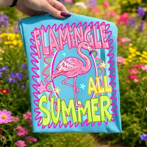 Puede incluir: Una camiseta azul claro con una ilustraci&oacute;n de un flamenco rosa y las palabras "Flamingle All Summer" en un dise&ntilde;o colorido. El flamenco est&aacute; de pie sobre una pata, rodeado de estrellas y un borde ondulado.
