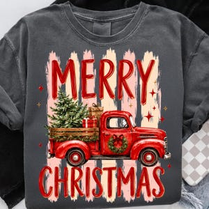 Może przedstawiać: Ciemnoszary t-shirt z grafiką czerwonego zabytkowego pickupa przewożącego choinkę i prezenty. Napis MERRY CHRISTMAS jest nadrukowany na czerwono nad i pod ciężar&oacute;wką, na tle pionowych pociągnięć pędzla.