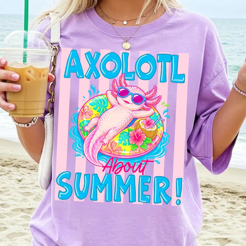 Peut inclure: T-shirt lilas avec un graphisme d'axolotl portant des lunettes de soleil et flottant sur un bou&eacute;e. Le texte "AXOLOTL About SUMMER!" est imprim&eacute; en bleu. L'axolotl est rose avec des accents roses et bleus.