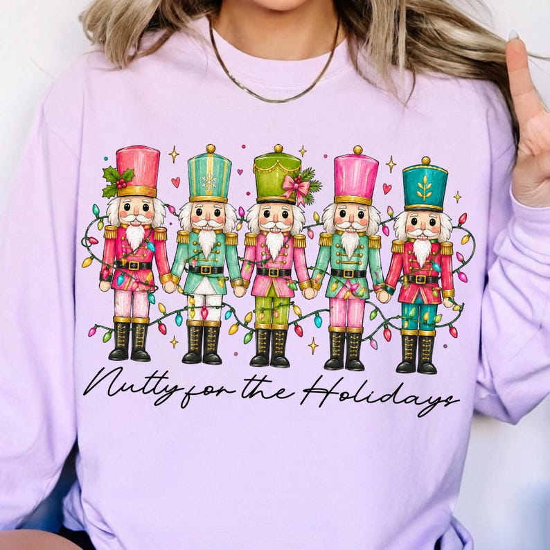 Op de afbeelding: Een lichtpaars sweatshirt met lange mouwen en een afbeelding van vijf notenkrakers in diverse pastelkleuren, verbonden door een snoer met gekleurde kerstverlichting. De tekst "Nutty for the Holidays" staat eronder in zwarte letters.