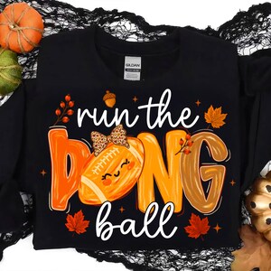 Puede incluir: Una camiseta negra de manga larga con el texto "run the ball" en letra blanca. El gr&aacute;fico incluye un bal&oacute;n de f&uacute;tbol americano con un lazo de estampado de leopardo, letras naranjas y marrones, hojas de oto&ntilde;o y peque&ntilde;as bayas.