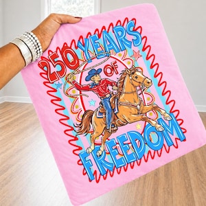 Puede incluir: Manta rosa con un vaquero a caballo con un lazo. El texto dice "250 YEARS OF FREEDOM" en rojo y azul. El vaquero lleva una camisa roja, vaqueros azules y un sombrero azul. La manta est&aacute; sujeta.